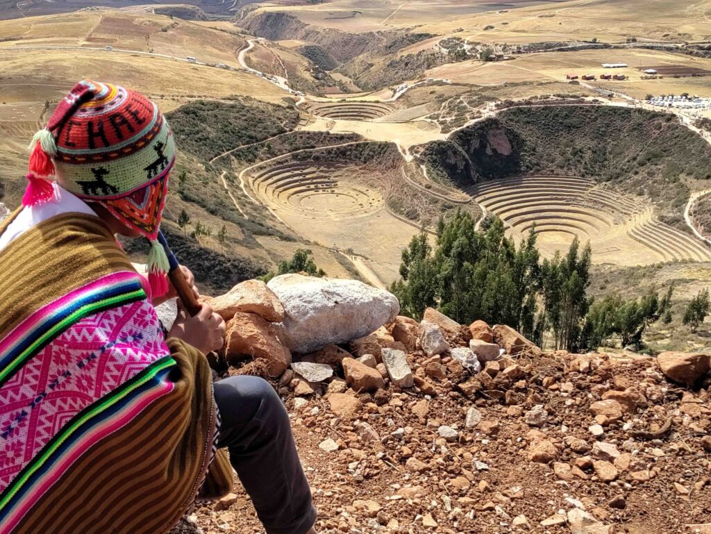A man dressed with traditional Peruvian hat and poncho looking out into the horizon as he's sat on a hill in peru