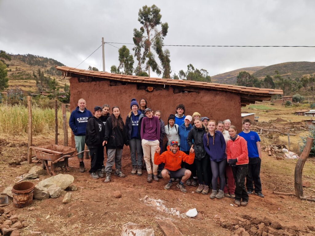 Group of student travellers standing in front of a hut they've built in Peru with the mountains behind them
