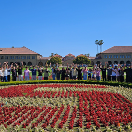 Big group of students standing around a flower bed displaying the Stanford University logo on a campus courtyard.