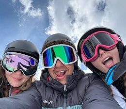 Three skiers who are taking a selfie from the ground up while wearing ski gear and snow goggles.