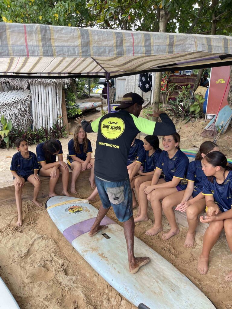 Group of students sitting in front of the coach while he teaches them about surfing standing on top of a board.