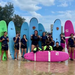 A group of students standing with their surf boards behind them while the coaches kneel in front to pose.